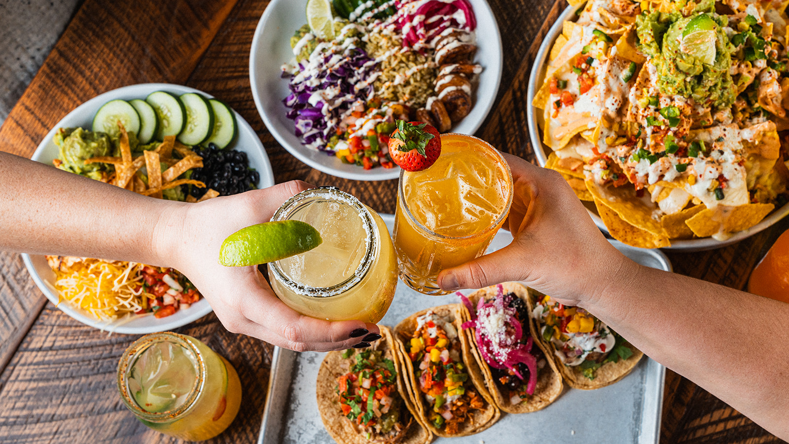 Two people clinking margaritas over a table of tacos and queso during happy hour at Social Cantina in Germantown, Nashville